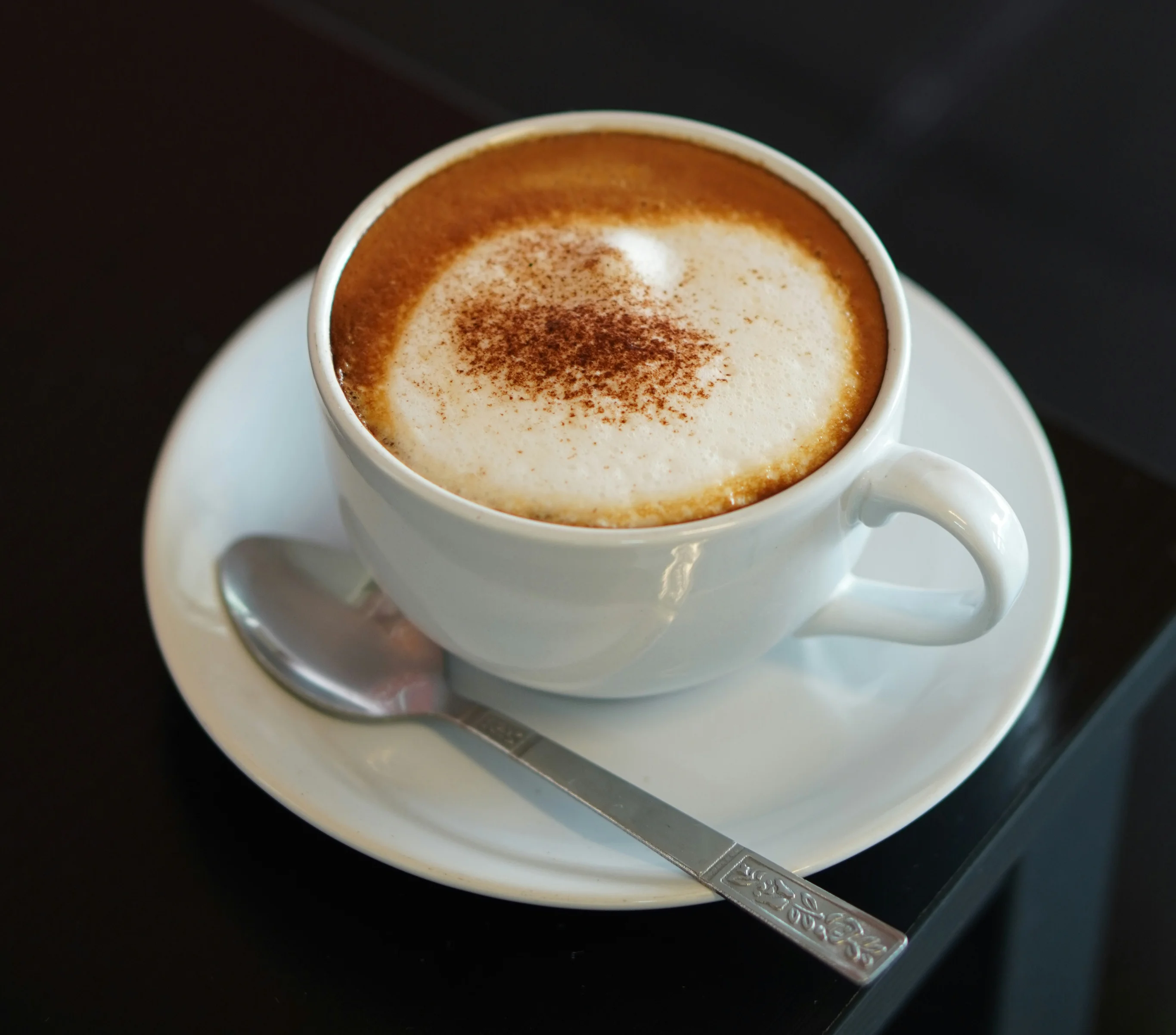 A close-up of a cappuccino in a white cup, topped with frothy milk and a sprinkle of cocoa powder, served on a saucer with a spoon.