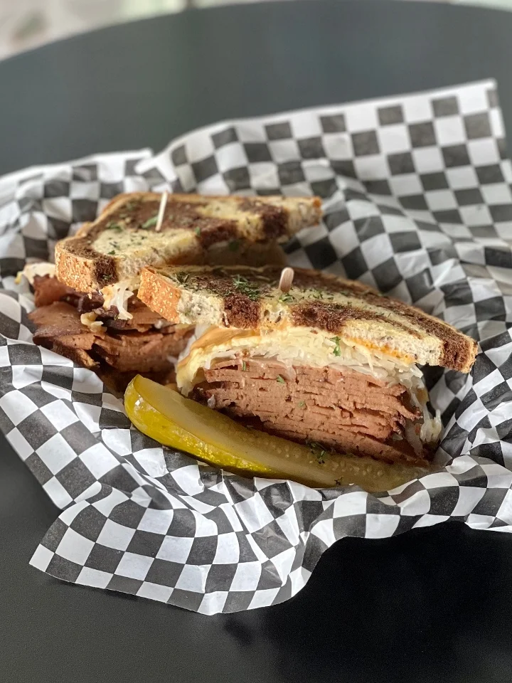 A sandwich filled with layers of meat served on toasted bread, accompanied by a pickle and placed in a checkered wrapper.