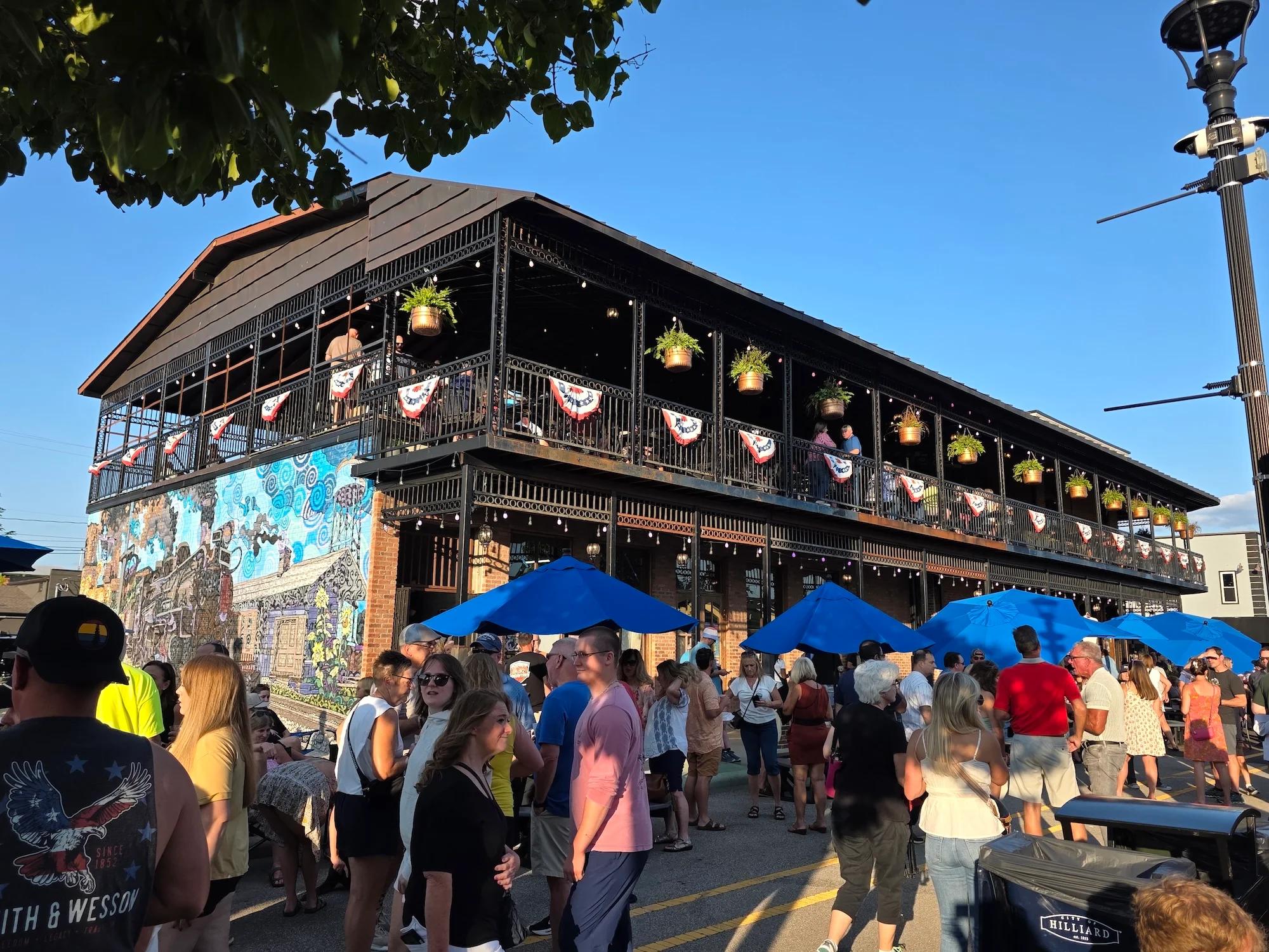 A vibrant outdoor scene featuring a busy venue with people gathered around, colorful decorations, and a mural on the side.