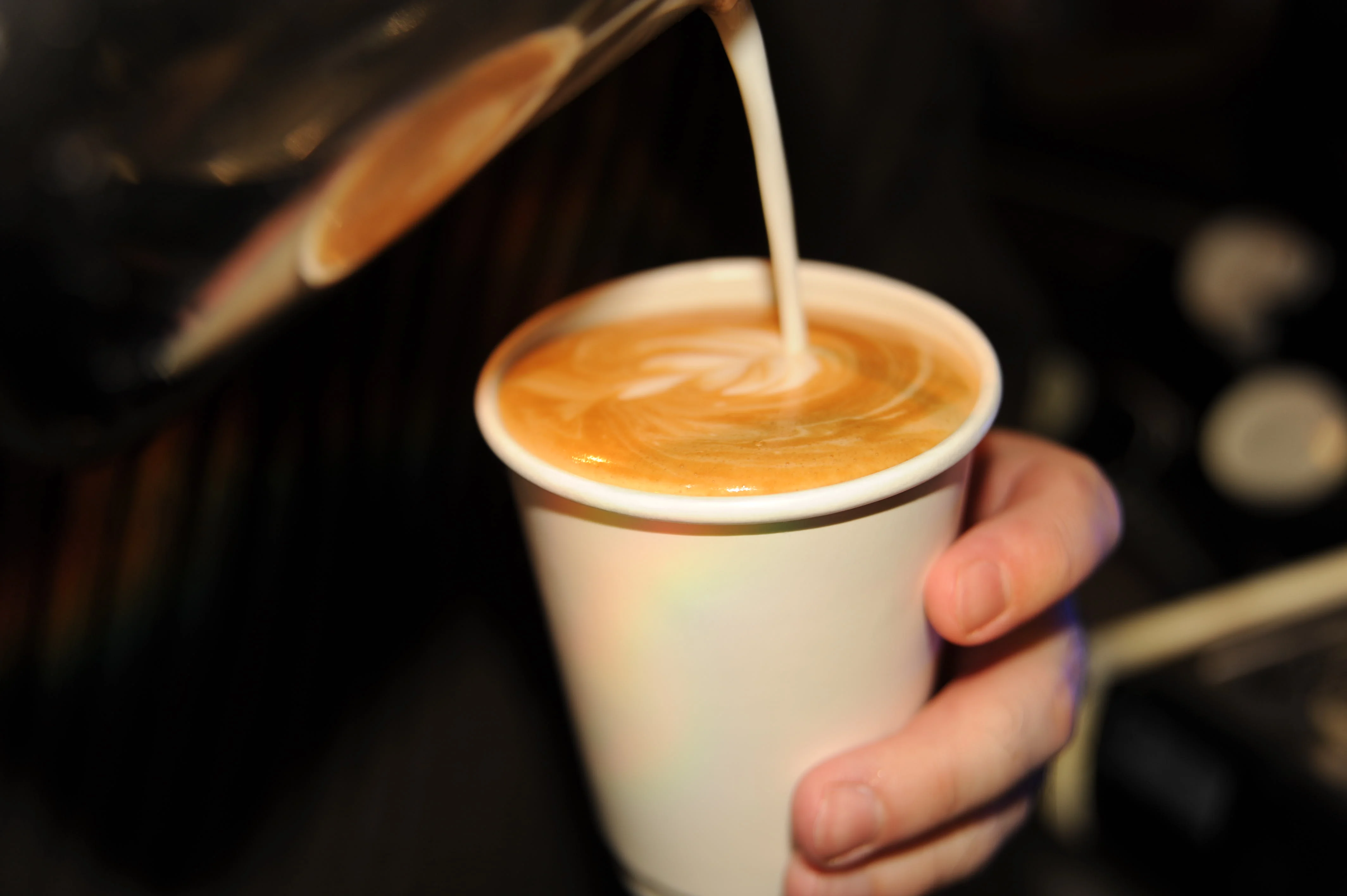 A close-up of coffee being poured from a metal pitcher into a white cup, creating an artistic swirl in the espresso.