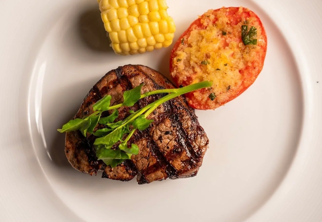 A grilled steak served with a piece of corn and a stuffed tomato half on a white plate.