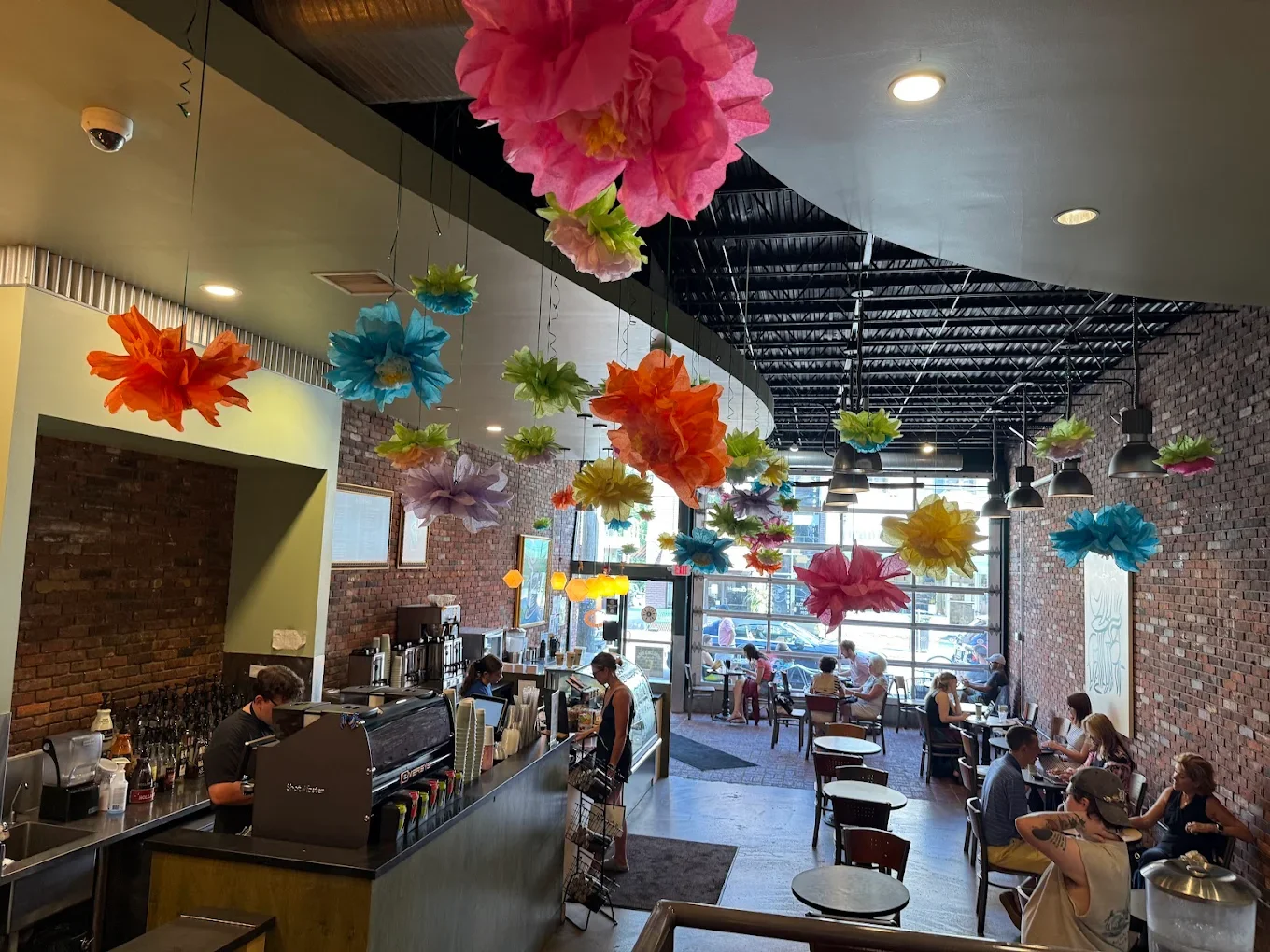 Colorful tissue paper flowers hang from the ceiling of a cozy café with exposed brick walls. Patrons enjoy coffee at tables, creating a lively, cheerful vibe.