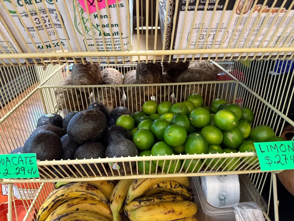 A display of fresh produce featuring avocados, limes, and bananas. The items are organized in a wire rack with signage indicating their prices.