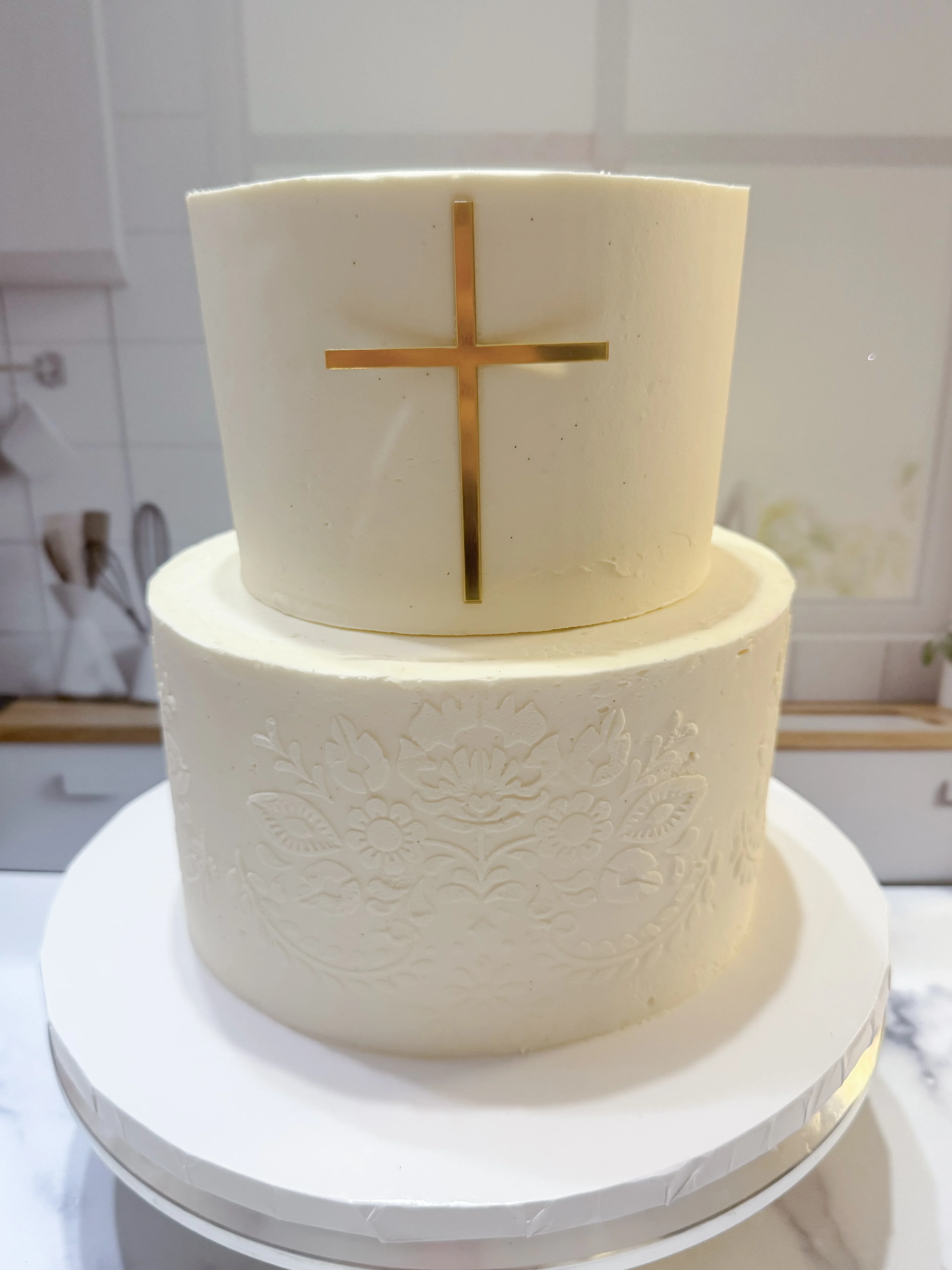 A two-tiered white cake adorned with a golden cross on the top layer. The surface features intricate floral patterns, and it sits on a round cake board.
