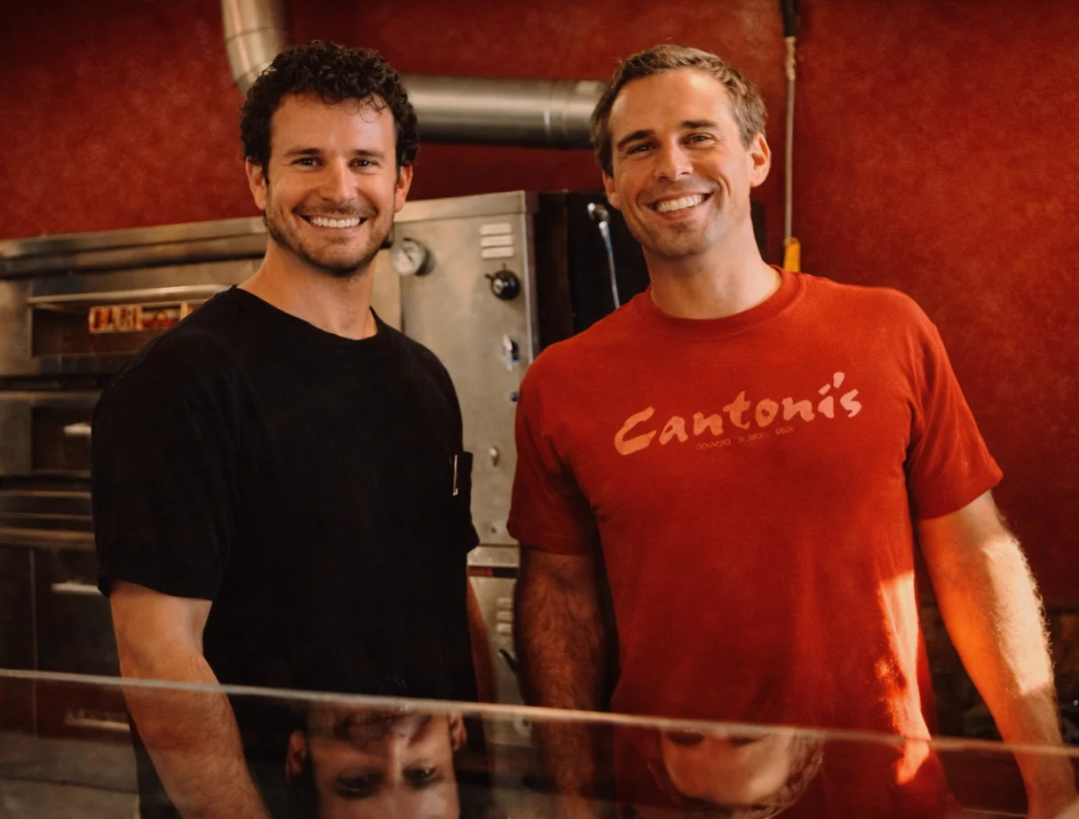 Two smiling individuals standing in a kitchen environment with a metal oven in the background.