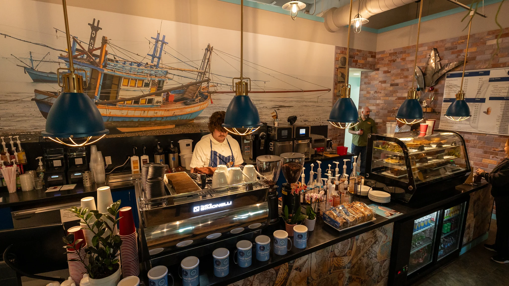 An interior view of a coffee shop featuring a barista preparing drinks, with a mural of fishing boats in the background.