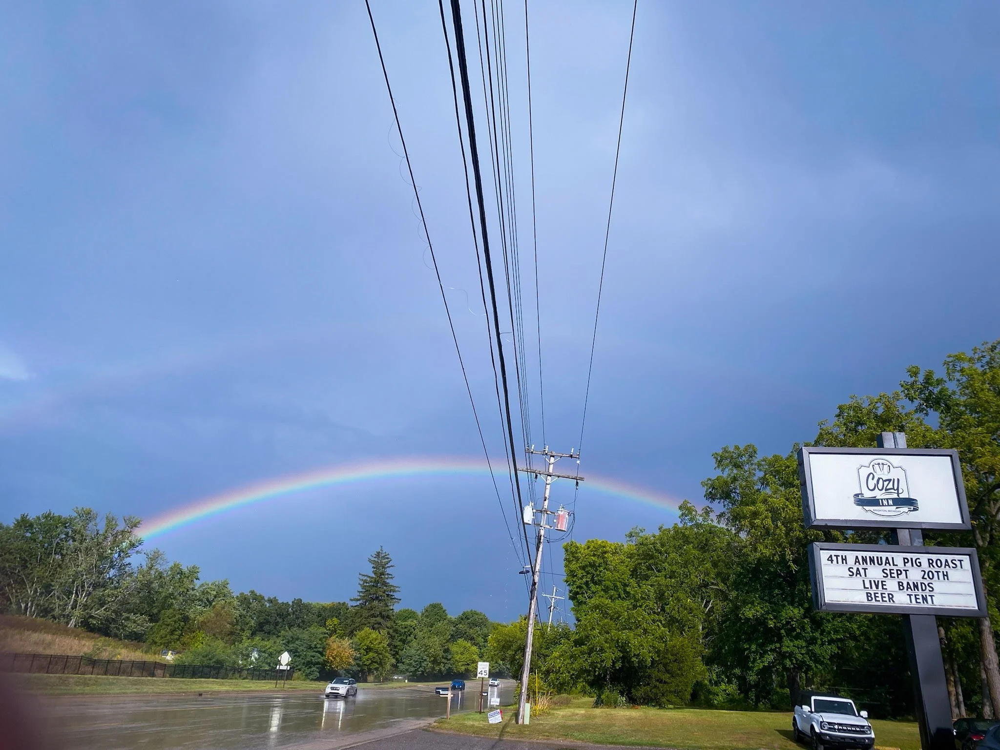 Rainbow over a road with a sign for a pig roast event at Cozy Inn.