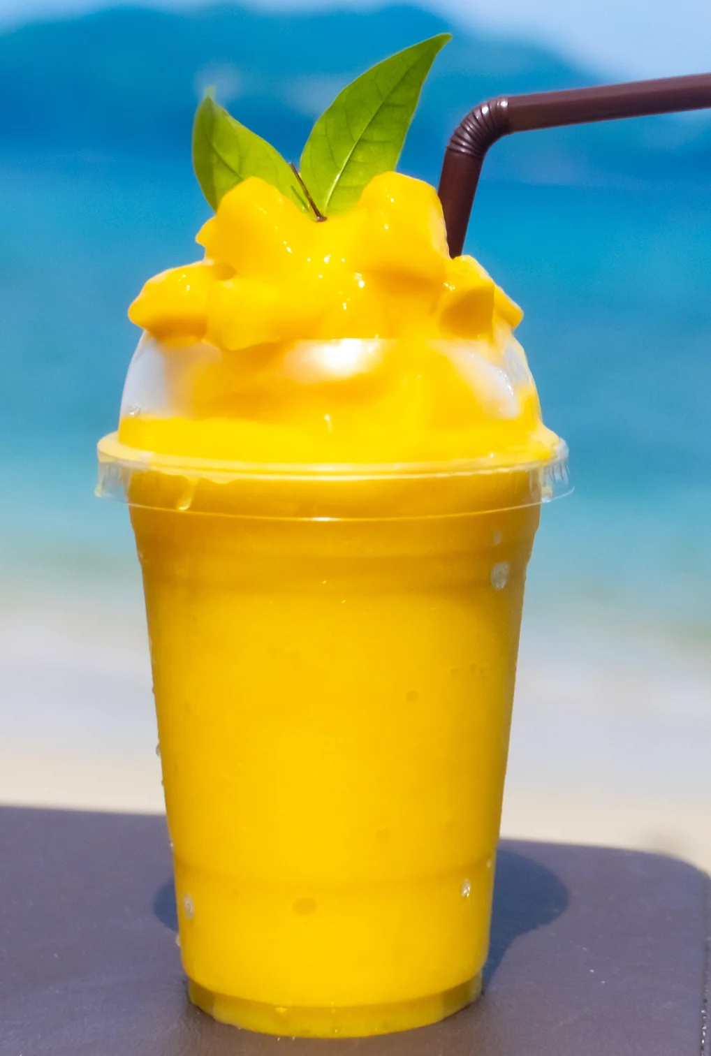 A vibrant mango smoothie topped with creamy whipped cream and a couple of green leaves, set against a beach backdrop.