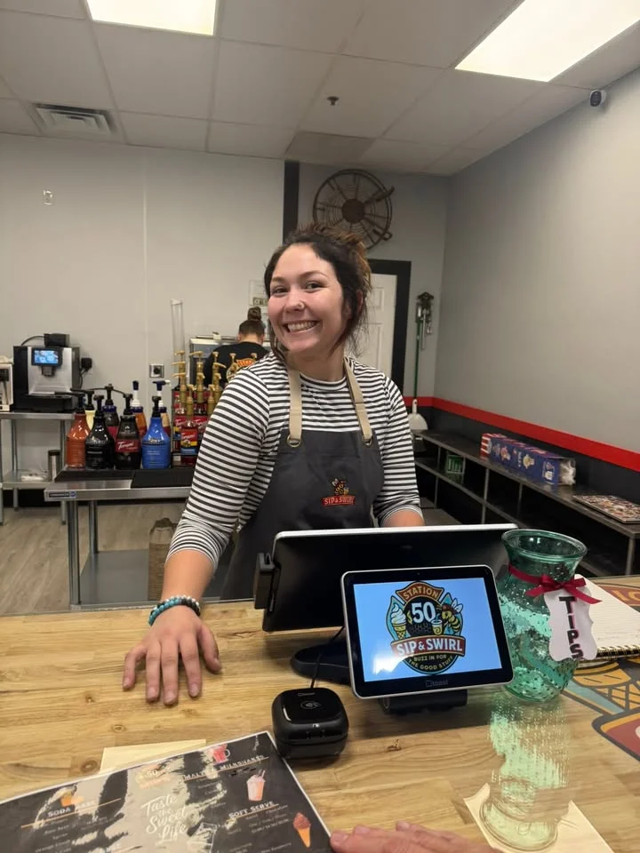 A cheerful staff member at a dessert shop stands behind the counter, ready to assist customers. The area features colorful beverage dispensers and a register.