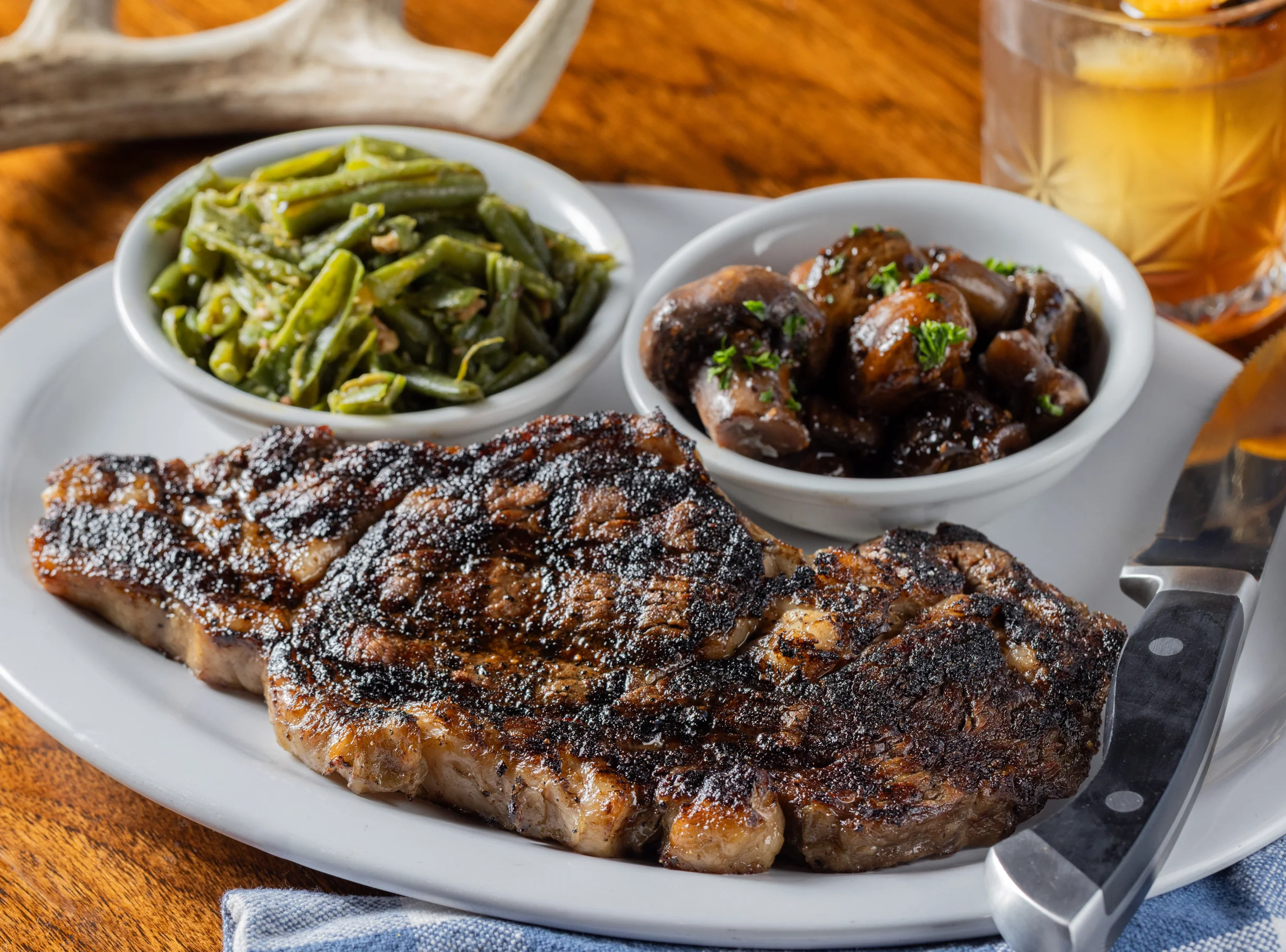Grilled steak with green beans, mushrooms, and a cocktail on a wooden table.