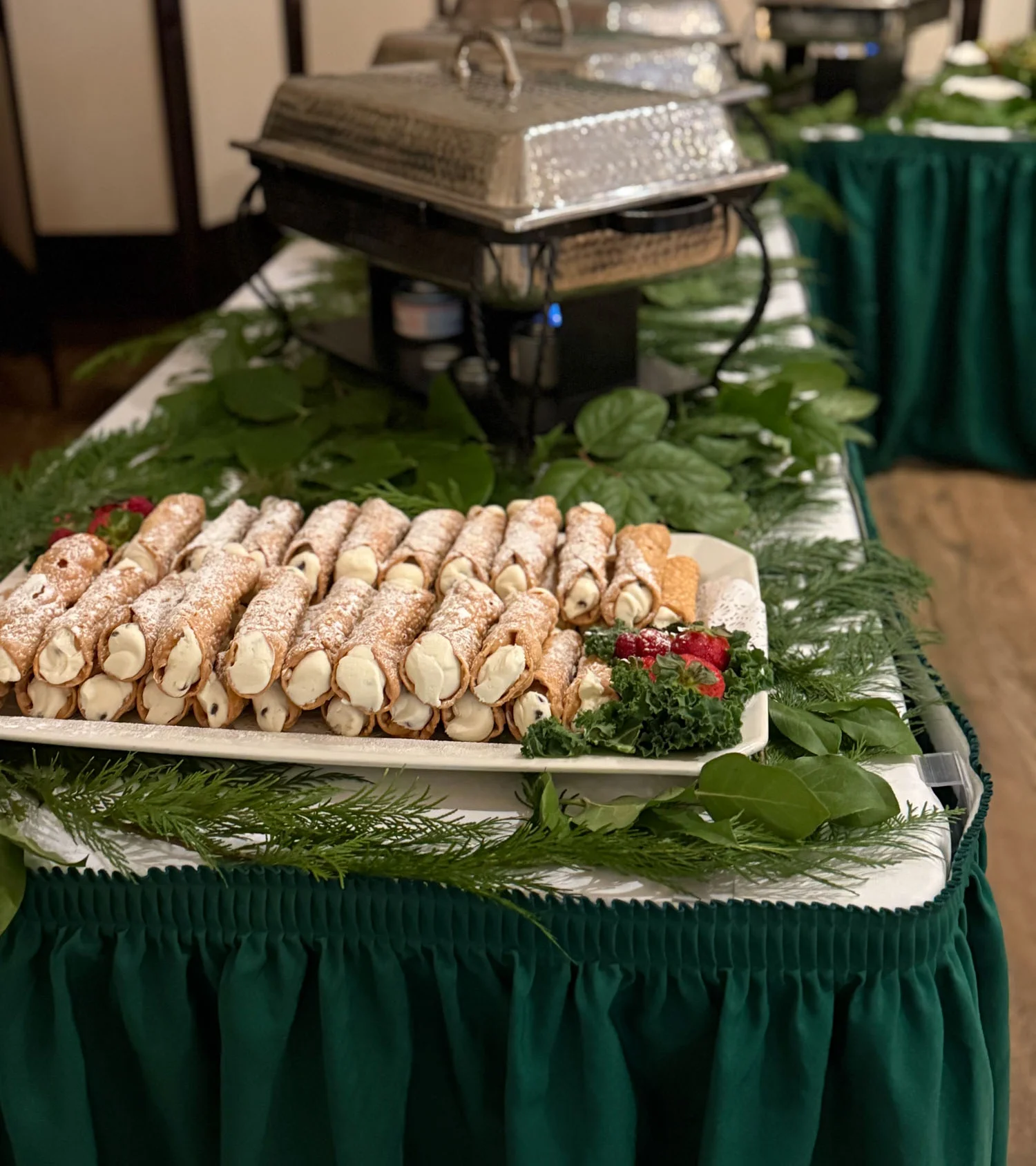 A tray of cannolis sit beautifully displayed on a long table. Additional chafing dishes can be seen lined up on the table behind the tray of desserts.