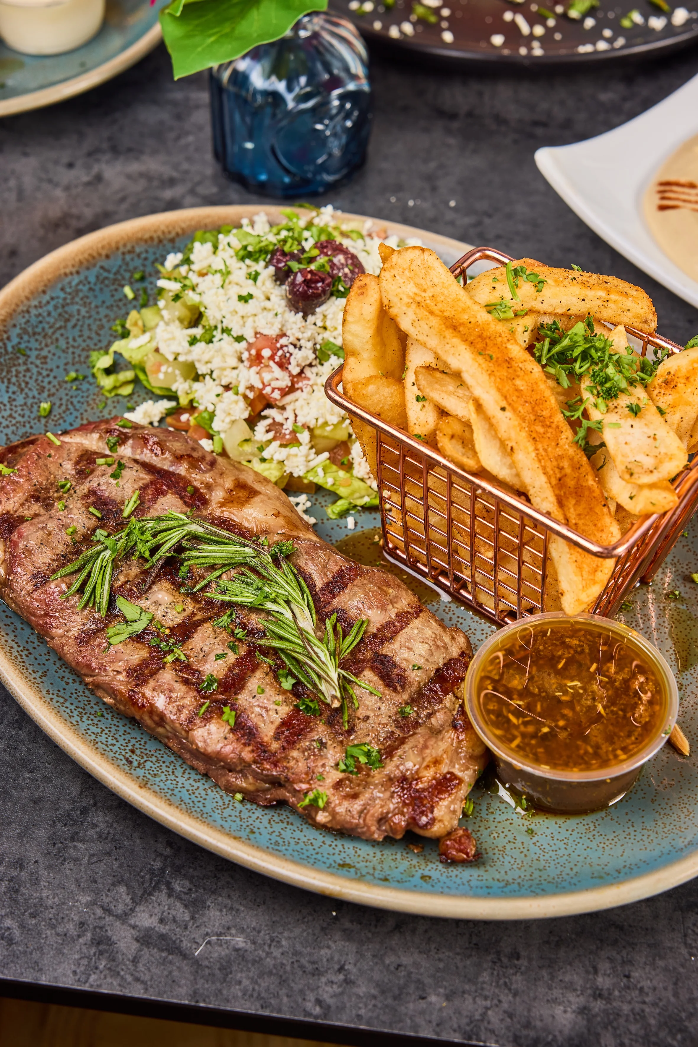 Grilled steak with rosemary, crinkle-cut fries in a basket, Greek salad, and dipping sauce on a blue speckled plate. Appealing and savory.