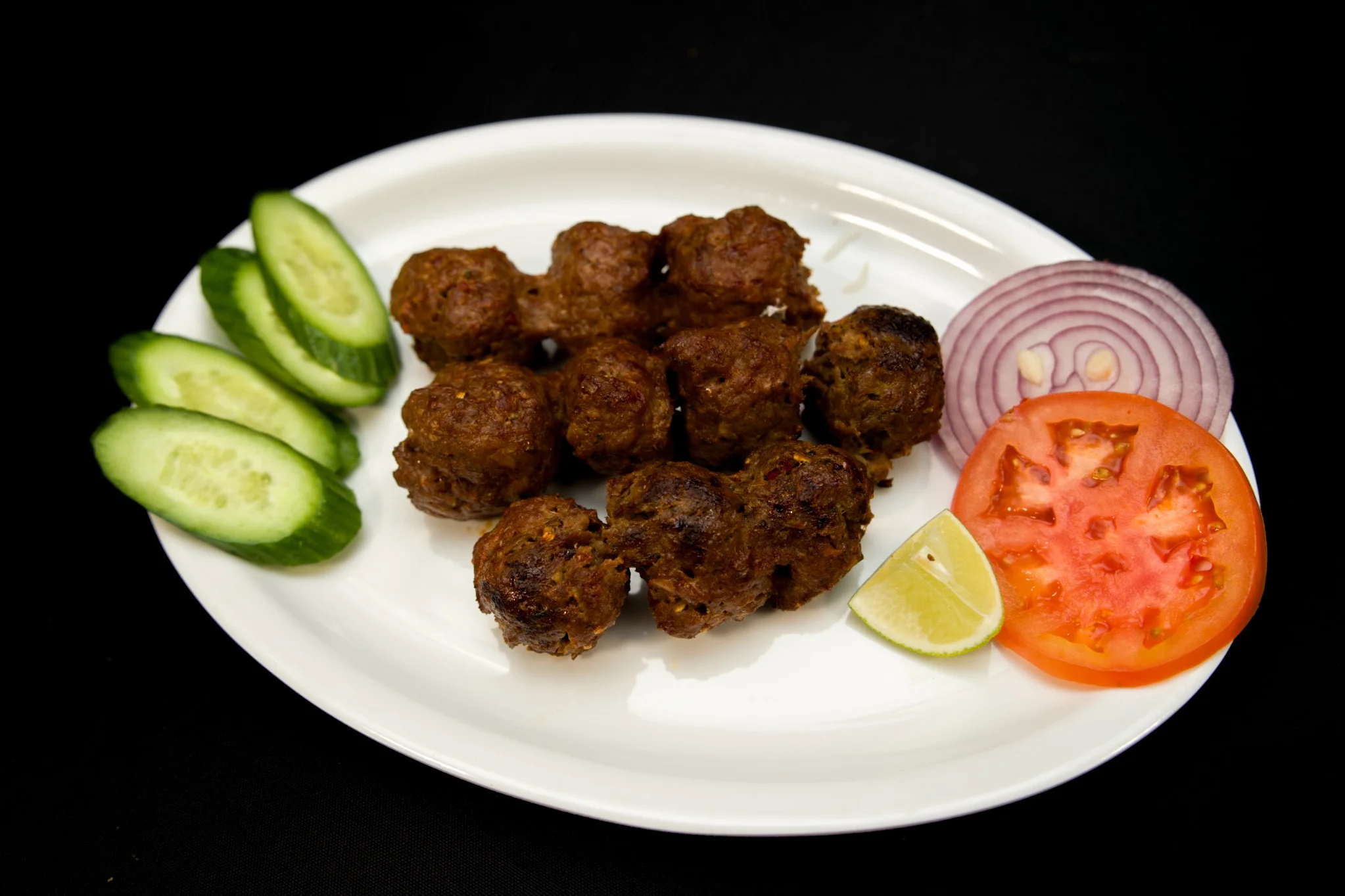 Plate of grilled Pakistani gola kabab served with sliced cucumber, tomato, red onion, and a lime wedge at Karachi BBQ Tonight.