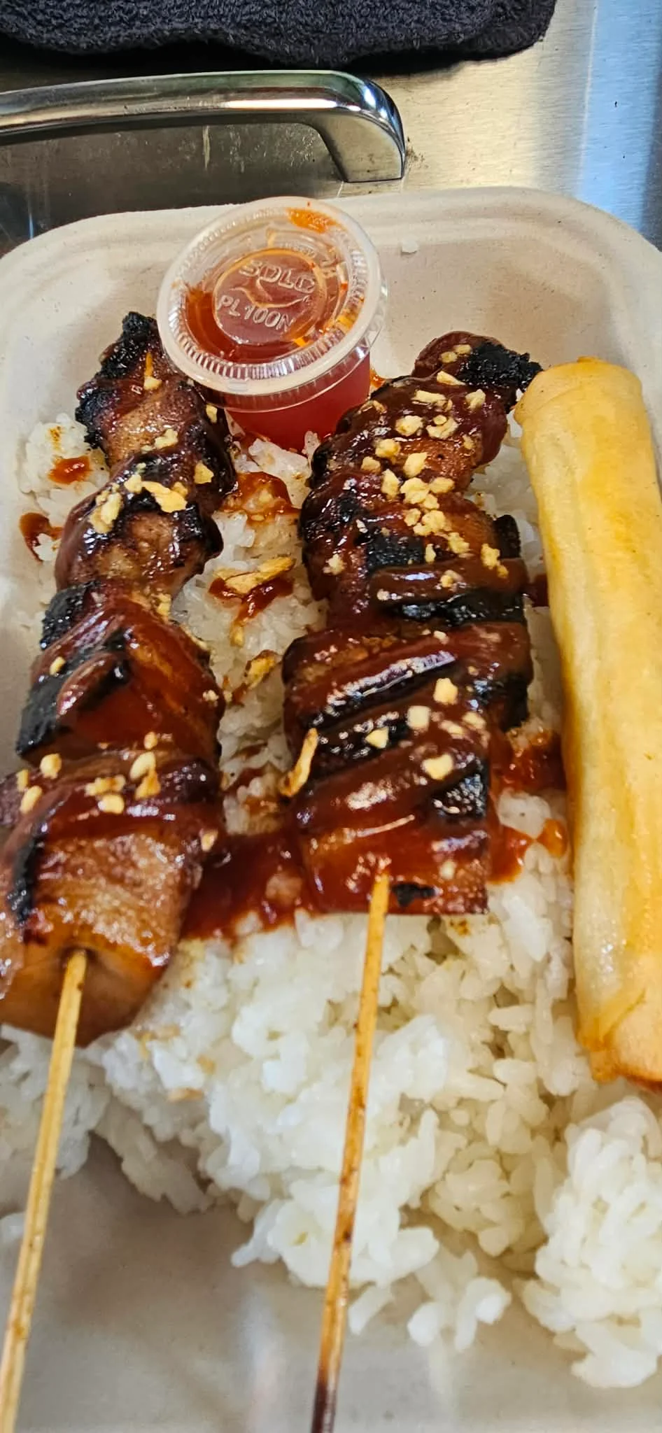 A serving of skewered meat satay drizzled with sauce, served with rice and a spring roll. The dish is garnished with crushed peanuts and accompanied by a small container of dipping sauce.