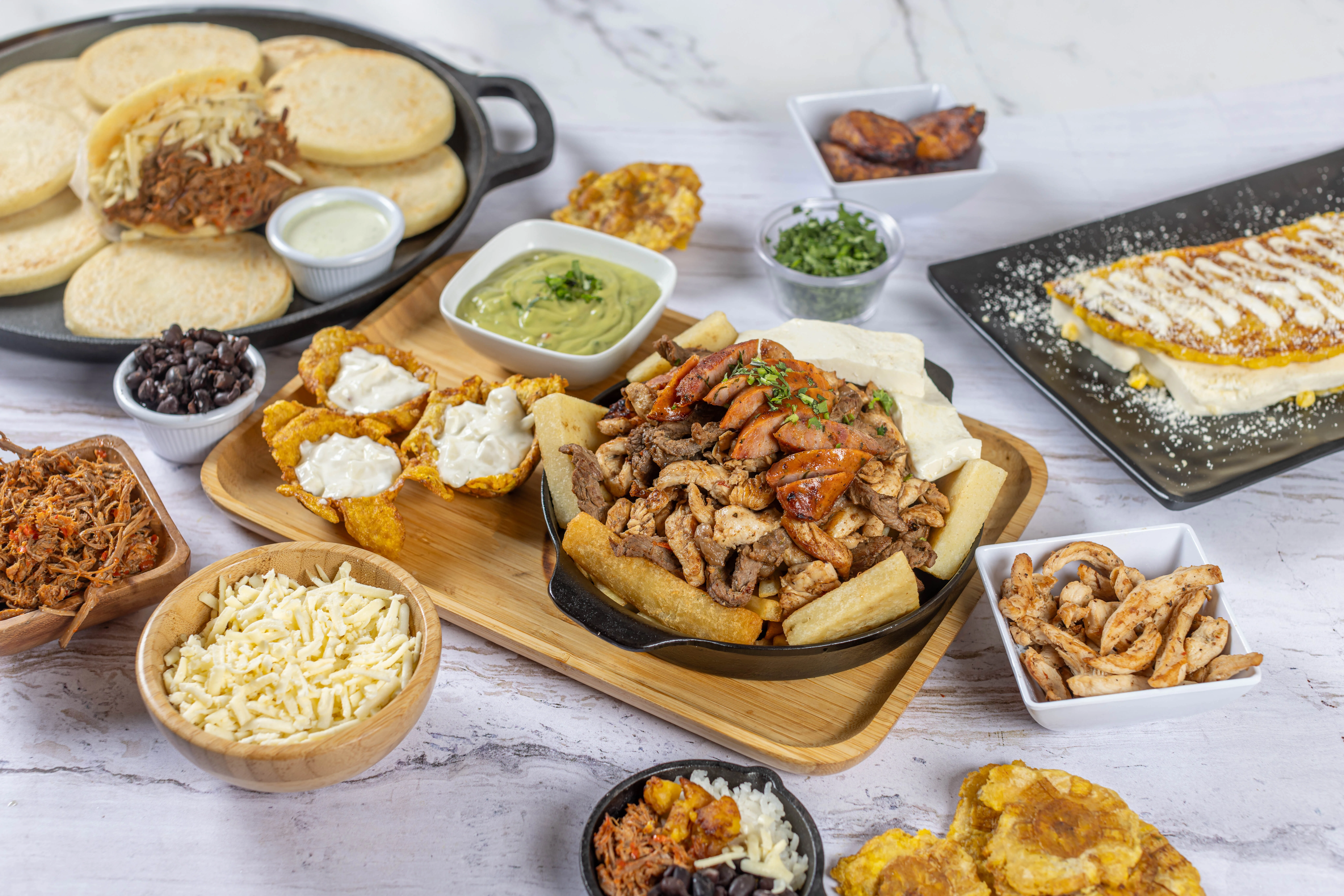 A large, family-style feast of Venezuelan food, featuring a central parrilla with grilled meats, surrounded by arepas, a cachapa, tostones, and bowls of cheese and beans.