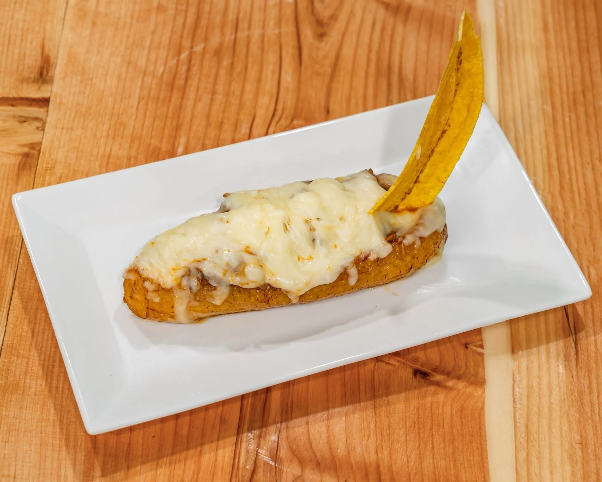 A traditional dish named 'Kanoa Montañesa,' featuring a ripe plantain sliced lengthwise and topped with melted cheese. The plantain is served on a rectangular white plate, garnished with a vertical plantain chip inserted into the cheese. The dish rests on a wooden surface.