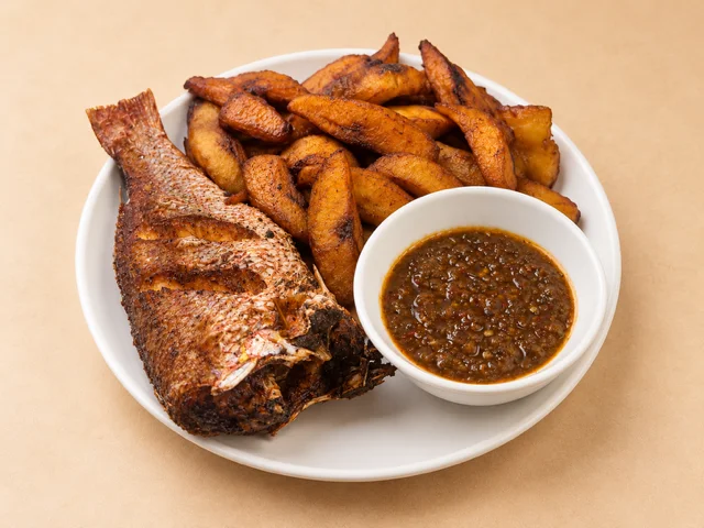 A plate featuring a whole grilled fish, served with crispy fried plantains and a bowl of spicy sauce.