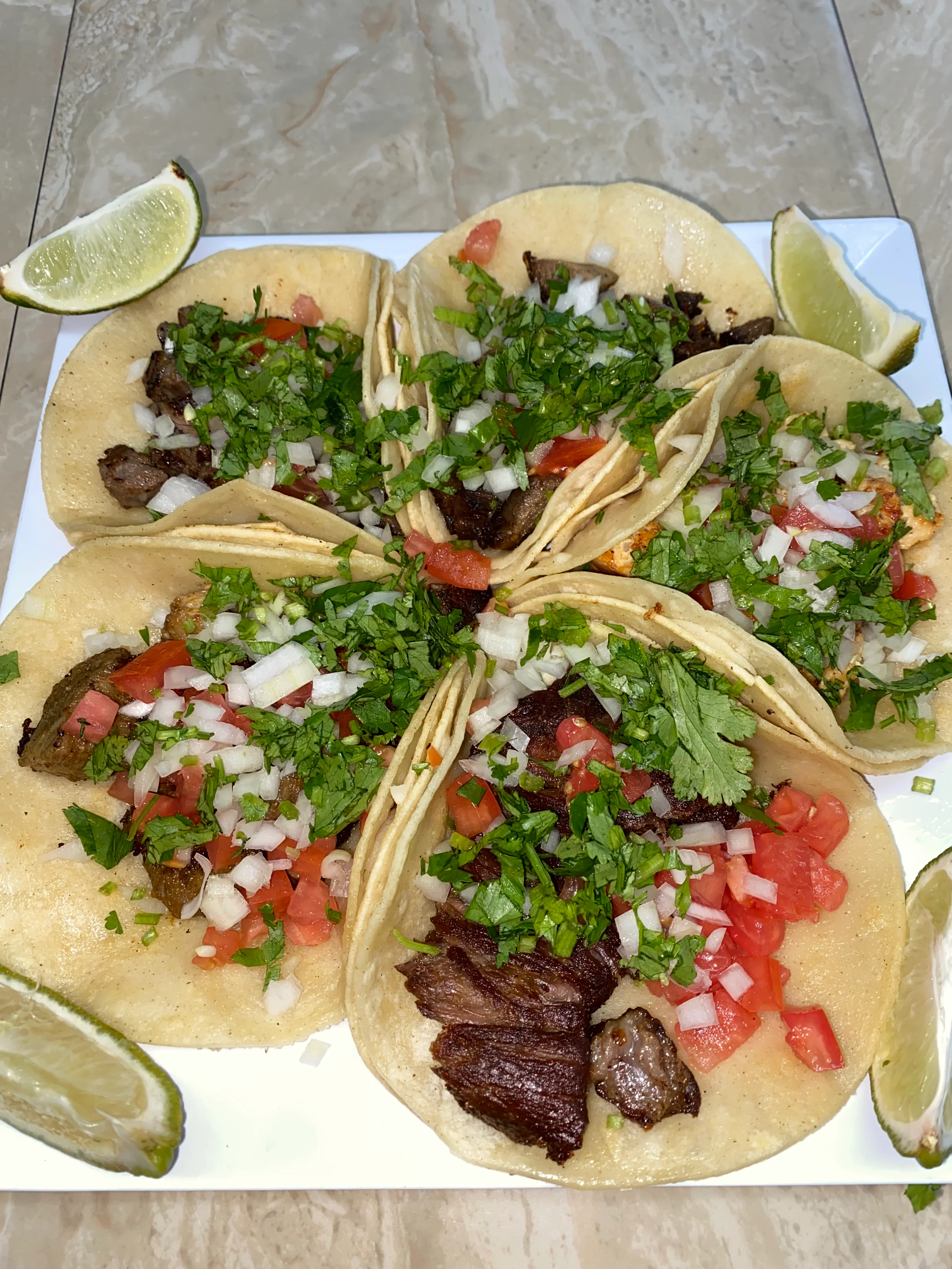 A platter of tacos filled with beef, topped with fresh cilantro, onions, and tomatoes, served with lime wedges.