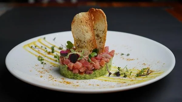 A gourmet dish featuring diced fish topped with herbs and served on a green puree, alongside crispy toast and decorative sauce lines.