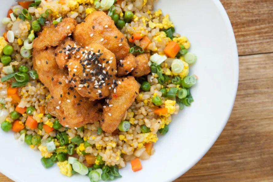 A hearty bowl featuring crispy fried pieces atop a colorful mix of grains, vegetables including peas, carrots, and green onions, and topped with sesame seeds.