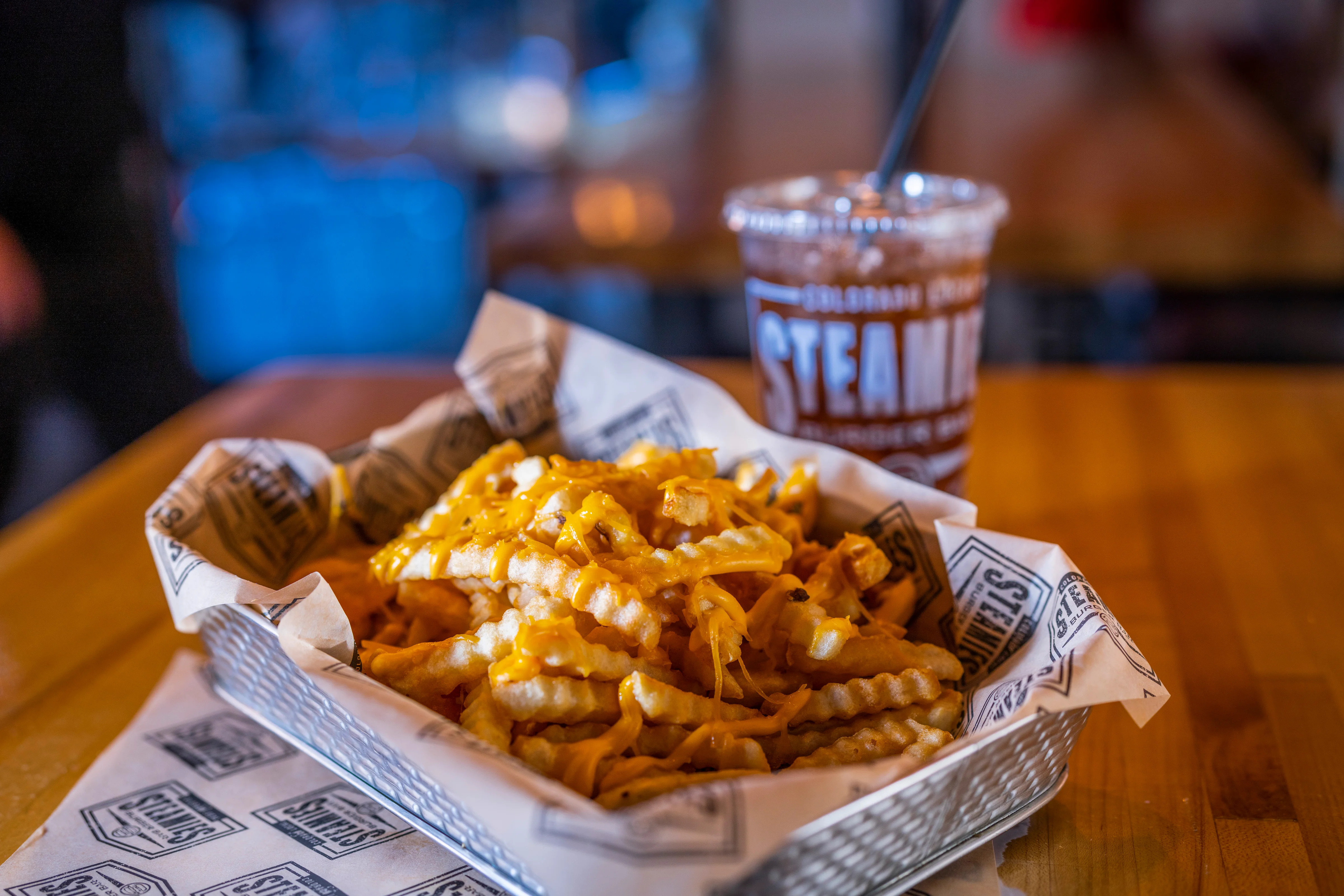 A basket of cheesy crinkle-cut fries accompanied by a drink, set on a wooden table.