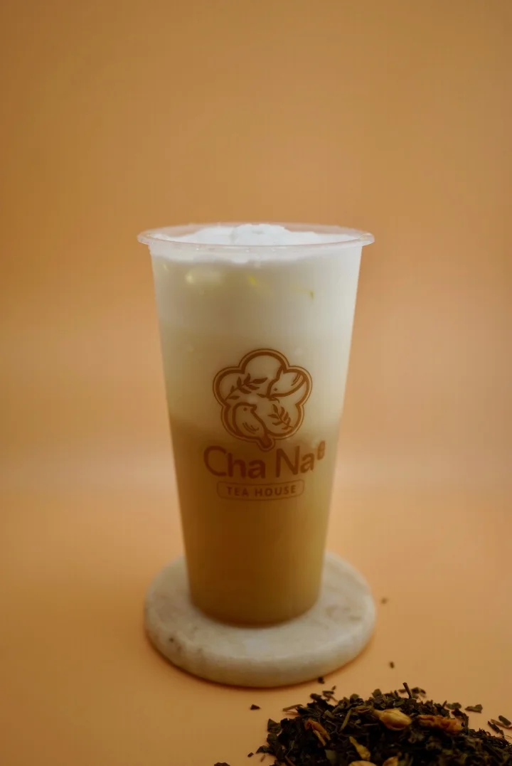A cup of tea with a creamy froth atop a light brown liquid, displayed on a stone base. The scene includes loose tea leaves on the side.