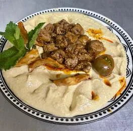 A bowl of hummus topped with spiced meat, garnished with fresh herbs and a drizzle of olive oil. The dish features decorative swirl patterns on the hummus.