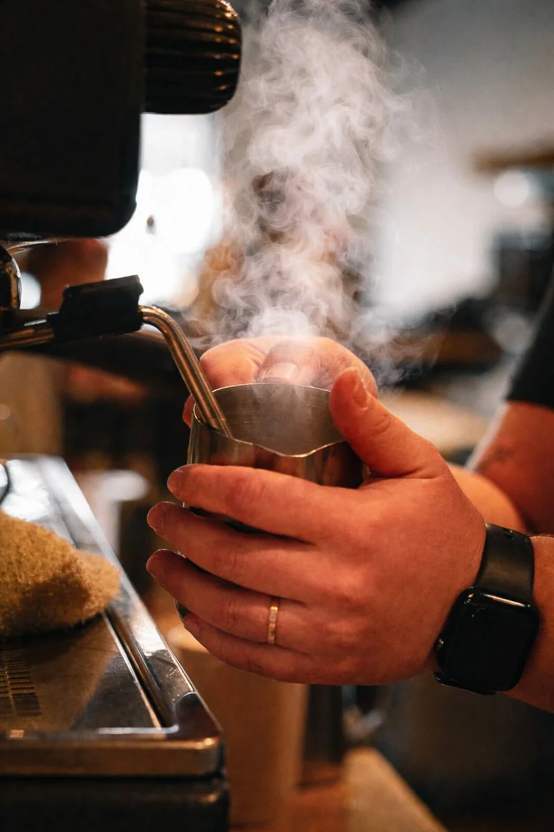 A barista is steaming milk in a metal jug, producing steam as they prepare a beverage. The setting suggests a coffee shop atmosphere.