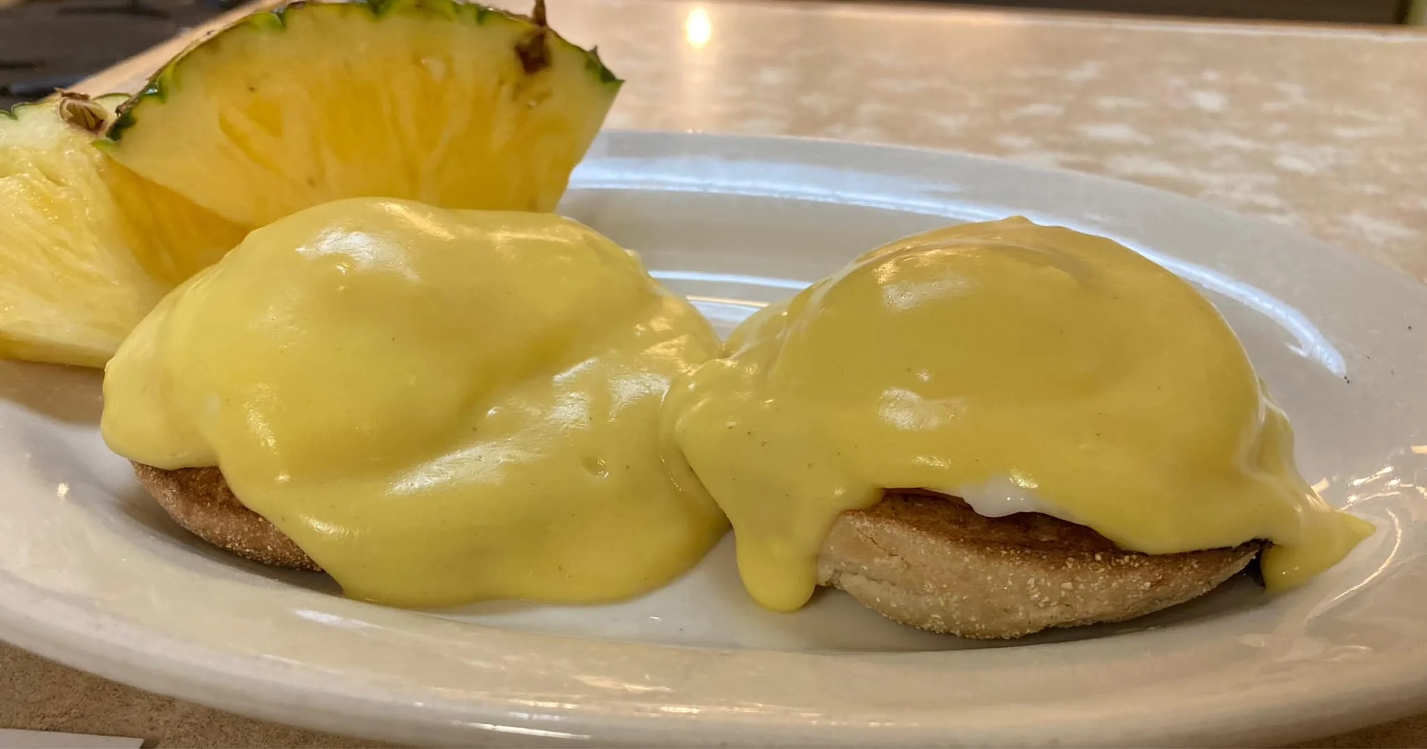 Two halves of an English muffin topped with creamy hollandaise sauce and served with a slice of pineapple.