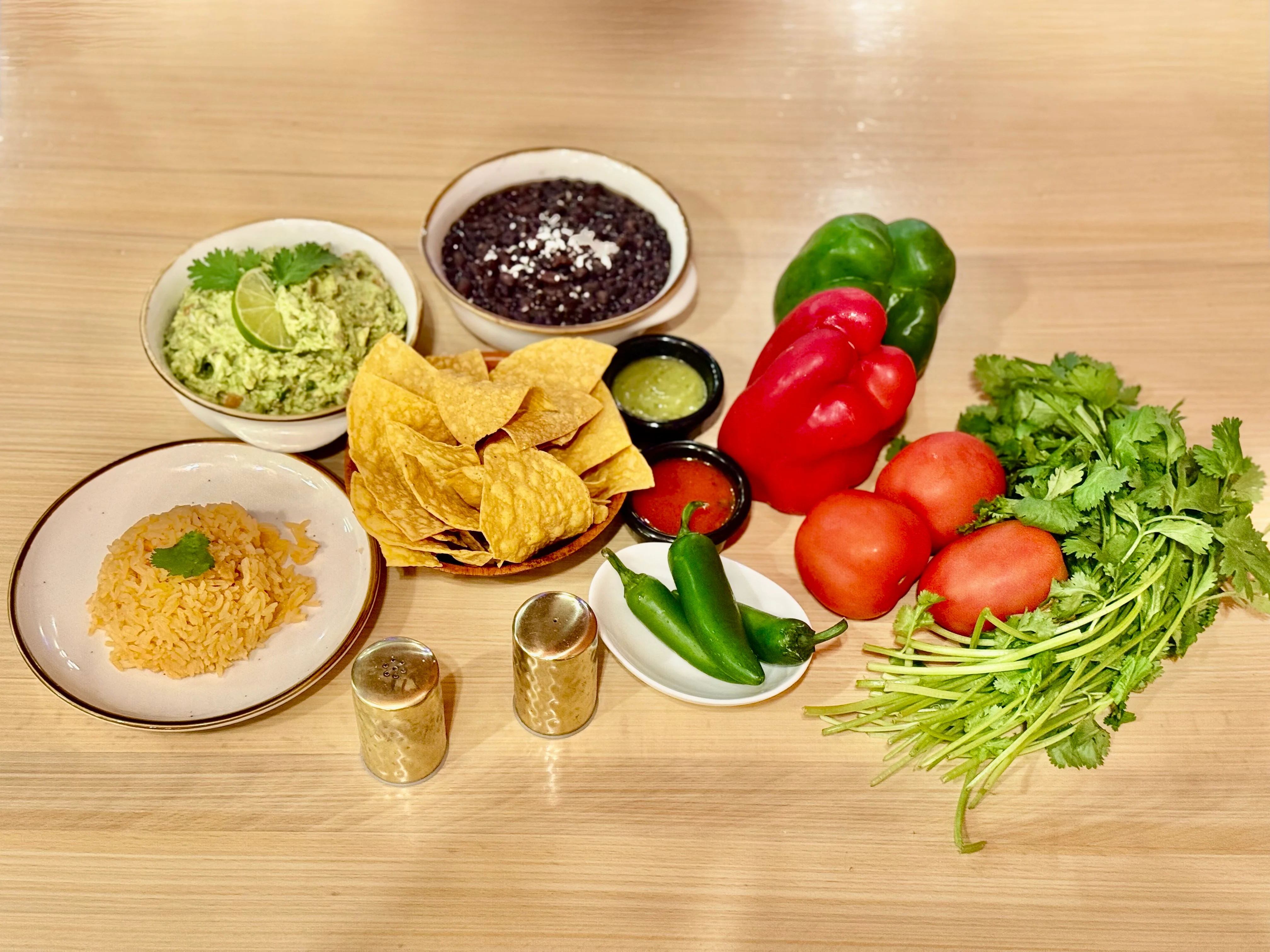 A colorful arrangement of various ingredients including guacamole, black beans, tortilla chips, rice, bell peppers, tomatoes, jalapeños, and fresh cilantro displayed on a wooden surface.