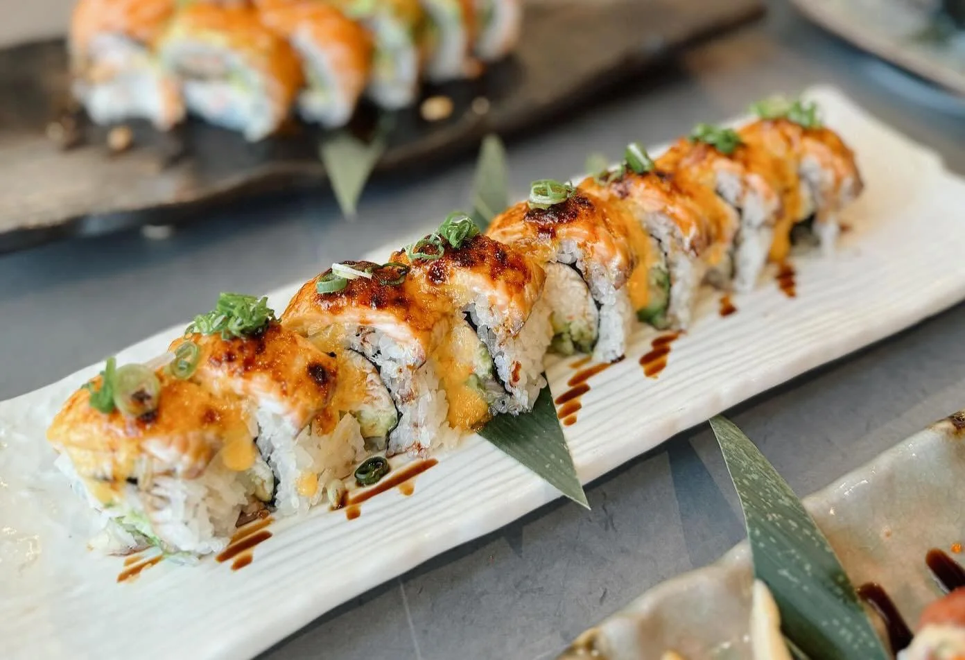 A stylized sushi roll topped with a golden glaze and garnished with green onions, served on a long white plate.