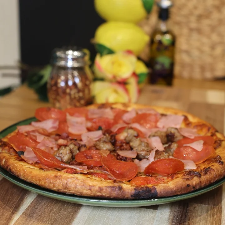 A savory pizza topped with pepperoni, ham, and sausage, served on a green plate with some fresh ingredients in the background.