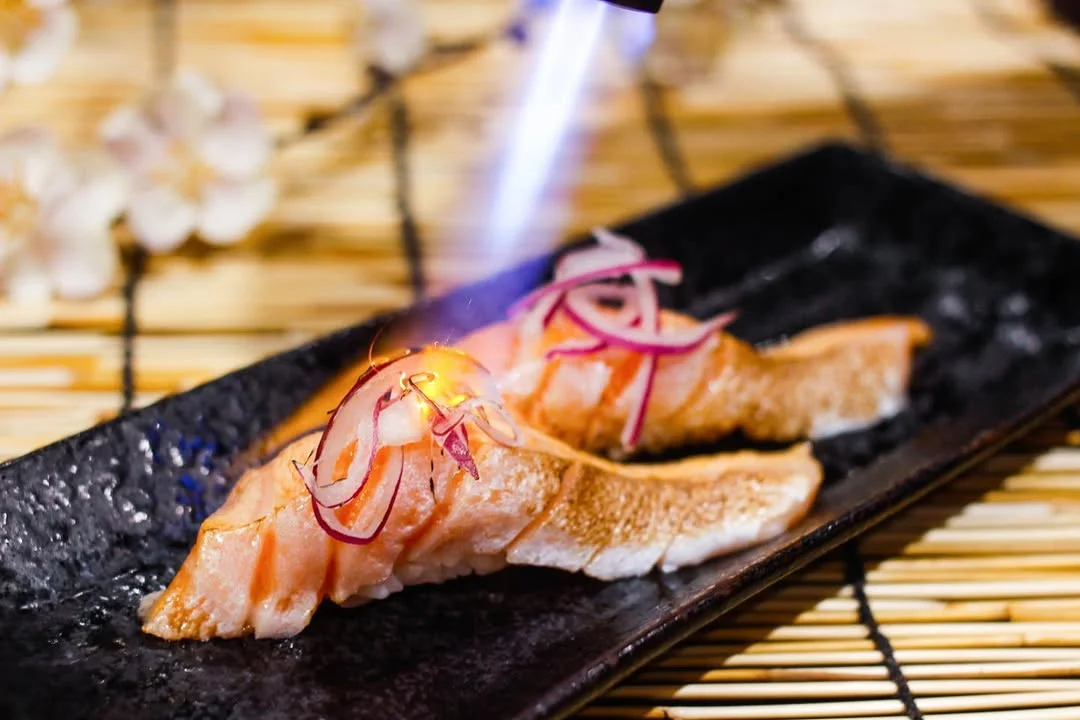 A plate of sushi with seared salmon topped with thinly sliced onions, being finished with a flamethrower for a smoky flavor.