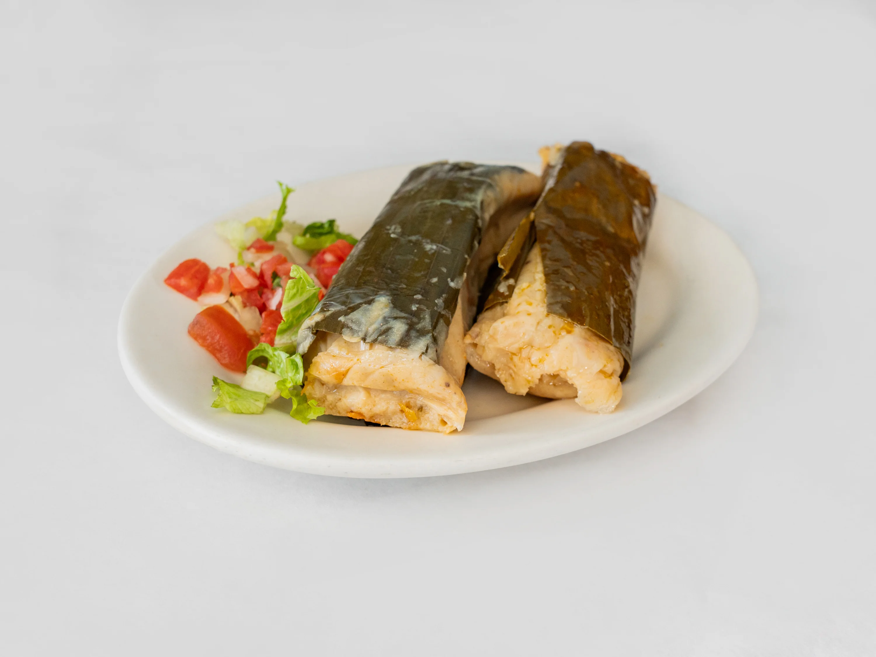 Two tamales wrapped in corn husks served with a fresh salad of chopped tomatoes and lettuce.